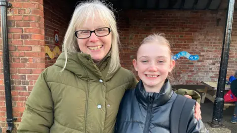 A mother and daughter stand close together smiling in the school playground. They both have blonde hair.