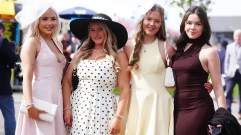 PA Media Four women stood in a line looking at the camera. One is wearing a pink dress and fascinator, one is wearing a black and white polka dot dress and black hat, a third is wearing a yellow dress and white fascinator and the fourth is wearing a brown dress.