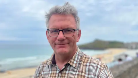 A man with grey hair and glasses wearing a brown and white checked shirt smiling slightly and looking directly at the camera with a beach in the background