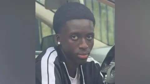 Baye Bireme Fall - a boy with short black hair, wearing a black and white striped tracksuit top, looking at the camera. He is sitting in a car.
