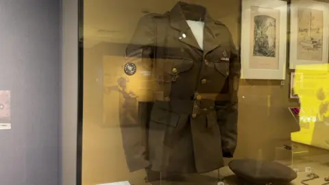 A khaki wartime uniform with brass buttons and a peaked collar behind a glass encasement.