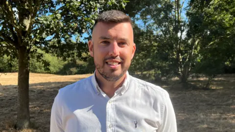 A man wearing a white shirt is looking at the camera smiling. He is standing in a field.