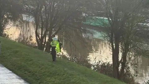 A police officer wearing a yellow high vis jacket and carrying a yellow high vis coat can be seen walking along grass next to a canal where trees and canal boats can be seen.