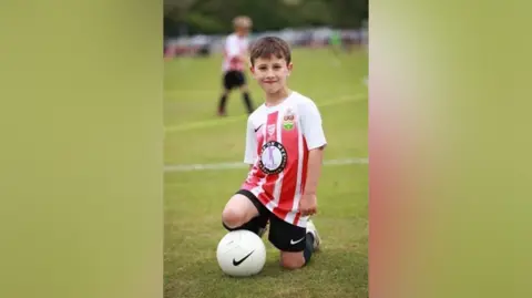 Family A boy kneels on a football pitch. He is wearing a red and white striped football shirt, and black shorts. One of his knees is resting on a white football with a black Nike logo on it.