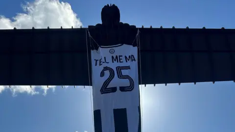 The back of the Angel of the North and the back of a Newcastle United top is hanging from the body. 'Tell me ma 25' is written on the back of the shirt.