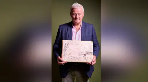 Roseberys Picture shows Stephen Hyde holding the book "Life and Landscape on the Norfolk Broads". Stephen wears a blue jacket and cream trousers. The book is large, he holds it in front of his body with both hands. The book cover is pale with gold lettering spelling out the title.