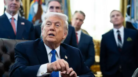 Donald Trump pictured sat behind his desk in the Oval Office. He wears a black suit jacket, white shirt, blue tie, and a pin of the US flag. Behind him stand four male officials in suits.