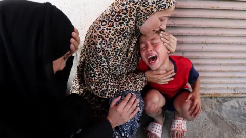A woman comforts a child at al-Aqsa Hospital in Deir al-Balah, after medics said at least 15 people were killed in an Israeli strike at a roundabout, in central Gaza (10 July 2025)