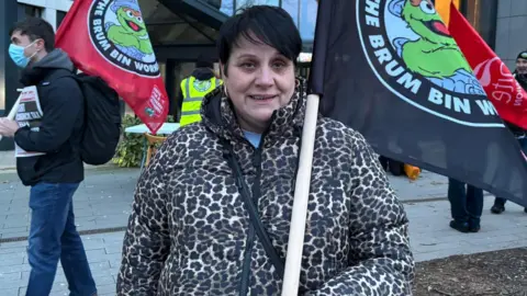 A woman with a leopard-print coat and holding a black flag with a cartoon creature and slogan on it, smiles as she attends a protest. Other protestors, one holding a flag, can be seen behind her.