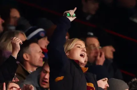 A young female fan cheers her team on, standing with a finger aloft, during the Bristol v Bath rugby game at Ashton Gate