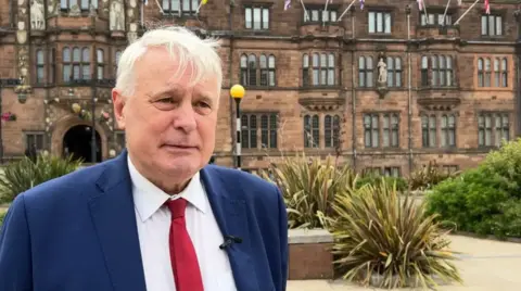 BBC A man with white hair in a blue suit jacket, white shirt and red tie, stood in front of a large red brick building with bushes behind him