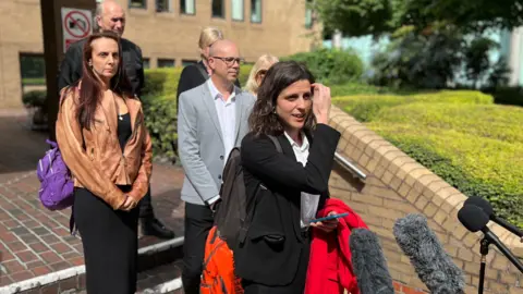 Five members of Donald Burgess's family stand in sunshine on the steps of Southwark Crown Court, as a member of their legal team, dressed in black, speaks to members of the press.
