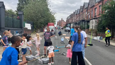 Ten children of various ages are playing in the street, which has been blocked off to traffic. In the foreground several children are making bubbles by dunking sticks and string in a bucket of liquid. And further up the street there are ramps for scooters and several more children playing football. 