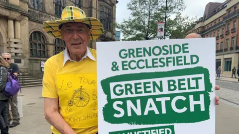BBC A man is wearing a big yellow checked hat with a bright yellow t-shirt with a bike design. He is holding a large green and white placard saying "green belt snatch" and is standing outside Sheffield town hall.
