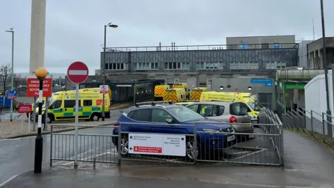 Ambulances and a couple of cars parked outside Derriford Hospital in Plymouth. It is a cloudy and wet day. The hospital building is grey.