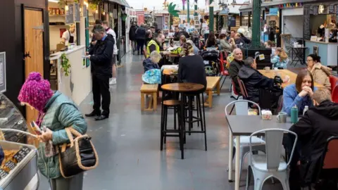 An indoor market lined with stalls on either side and tables and chairs in the centre. The markets is busy with people browsing stalls and eating.