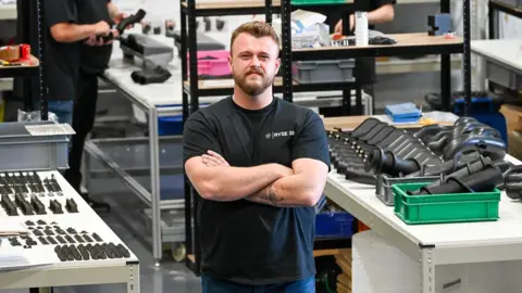 A man in a black t-shirt with his arms folded in a workshop surrounded by black components on tables. He has brown hair and a beard and a tattoo on his left arm. The bodies of others can be seen behind him, with parts in their hands.