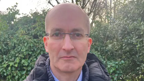 Professor Noel Purdy pictured outside, he is standing in front of a green bush. He has balding white hair and is wearing non framed glasses. He has a dark coat on.