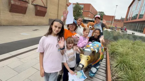 A group of children wave beside a colourful painted bear sculpture while holding trail maps in Scunthorpe town centre as part of the We're Going on a Bear Hunt art trail.