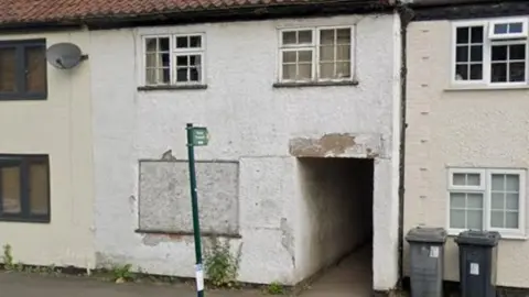 Google The white home has paint peeling off the walls and one of the windows is boarded up. 