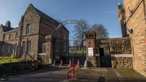 The outside gate at Cameron Barracks. An arched metal sign saying 'Cameron Barracks' sits above the main gate, which is attached to to the buildings by a brick wall. It's a clear sunny day. There are traffic cones outside the gate.