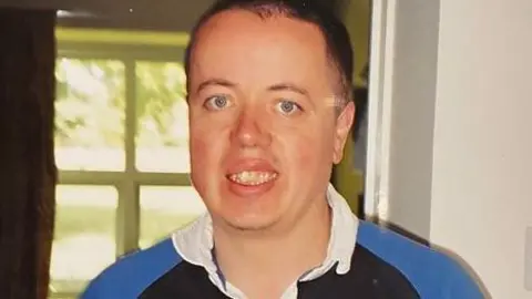 Family photo A young man with short dark hair smiling at the camera. He is wearing a blue rugby top.
