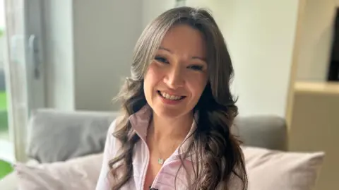 Rhiannan is sitting ona sofa and smiling at the camera. She has long dark wavy hair and is wearing a white zip-up top