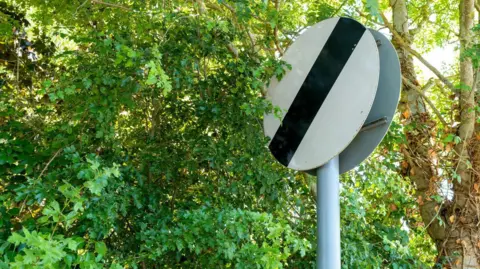Getty Images National speed limit road sign seen on a country road, the sign partially obscured by summer trees in foliage.