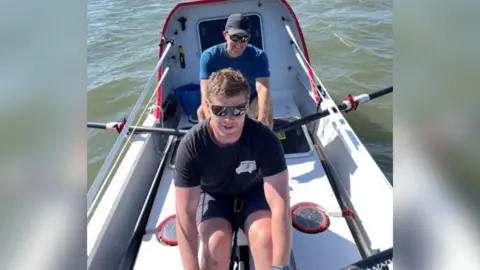 Henry and Damian pictured in a rowing boat, rowing through the water. Henry is sitting in the front and is wearing a black T-shirt and black shorts. Damian is sitting behind him wearing a blue T-shirt and a grey cap. They're both wearing sunglasses. 