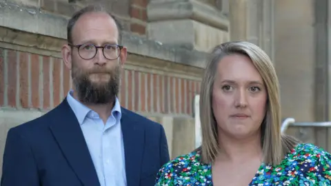 DJ McClaren/BBC Man with receding hair looks directly to camera. He is wearing round rimmed spectacles, a smart navy blue suit and light blue shirt with no tie. To the right of picture is a woman with bobbed shoulder length blonde hair. She has a multi-coloured floral dress on. They are stood in front of a red brick and stone public building.