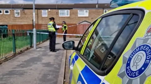 A police car with blue and yellow decor and a large Humberside Police badge is parked in a residential cul-de-sac. In the middle-distance, two police officers in black uniforms and hi-vis vests stand on either side of a cordon stretched across the street. In the background is a row of mid 20th-Century terraced houses.