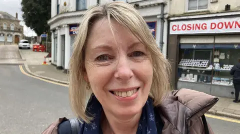 Susan Ashton stands in a street, smiling at the camera. She has a blonde bob haircut and is wearing a brown puffer jacket and blue floral shirt.