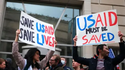 People stand in front of the US Agency for International Development (USAID) with placards saying "Save USAID, Save Lives"