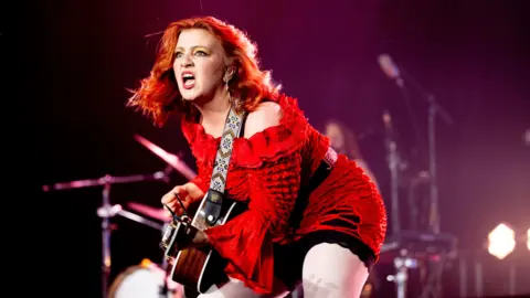CMAT, a woman with auburn hair plays a guitar on stage. She is wearing a red dress. Behind her out of focus, a drummer can be seen playing.