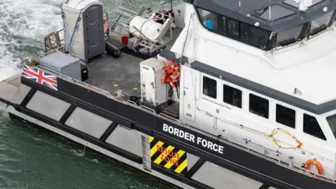 A Border Force vessel leaves the Port of Dover. The close-up photograph shows the equipment on the boat, with a Border Force sign, a Union flag and a "Rescue Zone" sign on the side. The water is choppy and white behind the vessel.