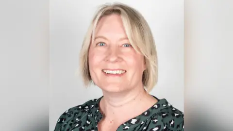 Hayley Douglas is smiling at the camera. She has blonde hair in bob style, blue eyes and is wearing a green, black and white dotted blouse. Behind her is a plain white background. 