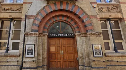 Google The Google image shows the two big wooden doors with a black sign atop for the Corn Exchange. There are two framed posters either side of the entrance against the cream coloured bricks and railings on both sides of the building leading up to the double doors. There are carvings in the upper level of the building, beneath two sets of windows, showing what looks to be agricultural workers and a horse and plough. 