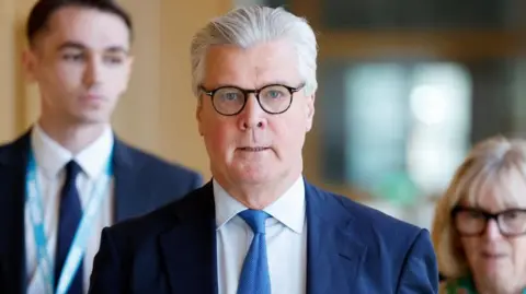Malcolm Offord, who has glasses and grey hair swept back, front on to the camera while wearing a business suit 
