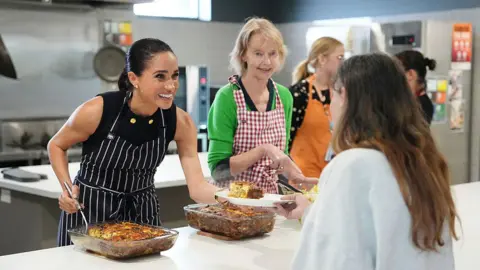 Getty Images Meghan, the Duchess of Sussex, wearing an apron and serving lunch alongside other helpers in a kitchen