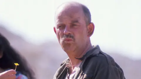 Julian Holloway in character as Paterson in Doctor Who. He has short cropped hair and a short moustache. He is frowning with pursed lips.