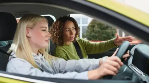 A driver and instructor sit in a car. They are both female and the driver's window is down giving a clearer image of them in the vehicle. The driver has blonde hair and is wearing a grey sweat top and has both hands on the wheel. The instructor has brown curly hair and is in a green top with one hand directing the driver.