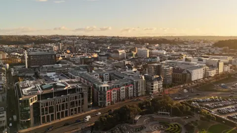 St Helier in evening light viewed from a drone. There is a park and a car park in the foreground and high rise buildings line the main road and stretch back in to the distance.