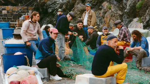 Ian Kingsnorth A scene from the film in which about 15 men are sitting on the quay with a rocky cliff behind them. They are acting as fishermen. Two of them are mending nets, some are sitting on upturned fish boxes and others are leaning against barrels of fishing line. Some are wearing yellow oilies and most of them are wearing caps or hats.