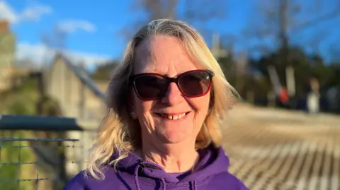 Clare has blonde shoulder length hair and is wearing brown sunglasses and a purple hoodie.