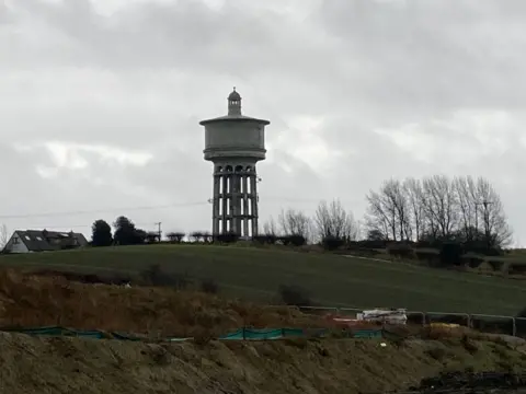 LDRS A concrete water tower in the distance surrounded by green fields and the roof of a house in the far left