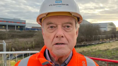 Colin Skellett, pictures while talking on site. He has light grey hair and light eyebrows, and is wearing a collared shirt with an orange high-vis jacket and a white hard hat with the words 'YTL Construction UK' on it. 