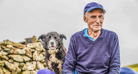 Stephen Wilson A man with white hair and a blue cap sitting next to a wet sheepdog and pile of stones. He is looking to the side wearing a blue jumper. The dog is looking into the camera.