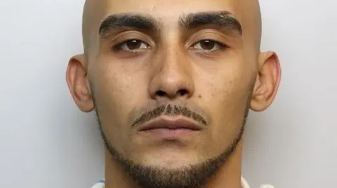 A bald man with a trimmed mustache, beard and thick eyebrows is posing for a mug shot in a police station.