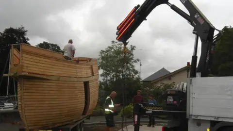 Men operate heavy lifting gear next to a large wooden boat. One of the men is in high vis. The sky is grey. 