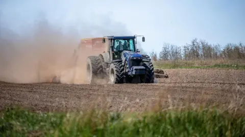 A large blue tractor driving on a field. There is a cloud of dust coming from the side of it and a red box behind it. The field is full of brown earth